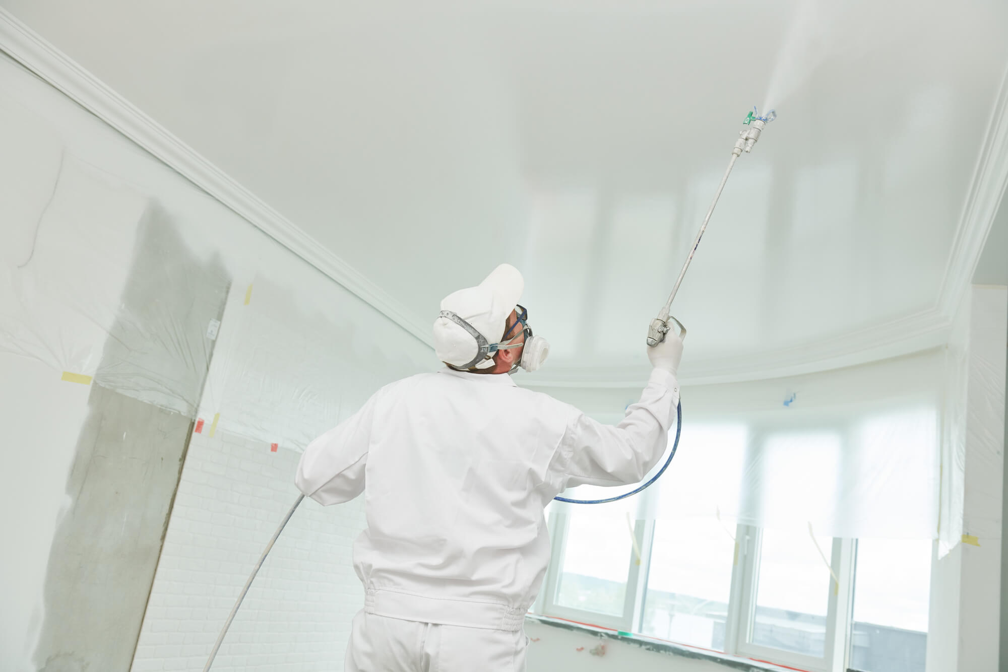 painter in white work suite spraying white ceiling in a room